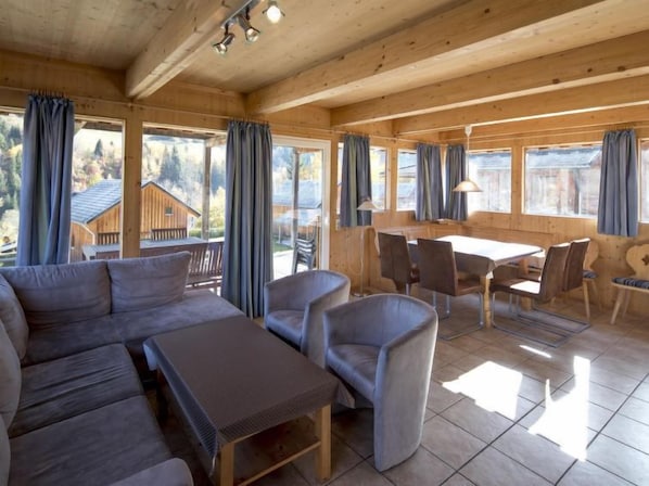 Un chalet en bois chaleureux, doté d’un canapé, de fauteuils et d’une table à manger. De grandes fenêtres, ornées de rideaux, offrent une vue sur les environs.