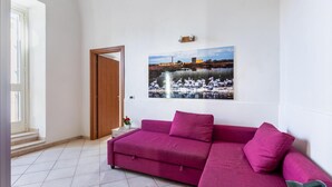 A living room with a purple sofa, a wall-mounted picture of a lake with birds, and a wooden door. - Ciuri Ciuri in Noto With 3 Bedrooms and 2 Bathrooms (Noto)