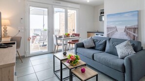 A modern living room with a grey sofa, a coffee table with flowers, and a painting of a beach. - Apartment SC H01 by Interhome (Mimizan)
