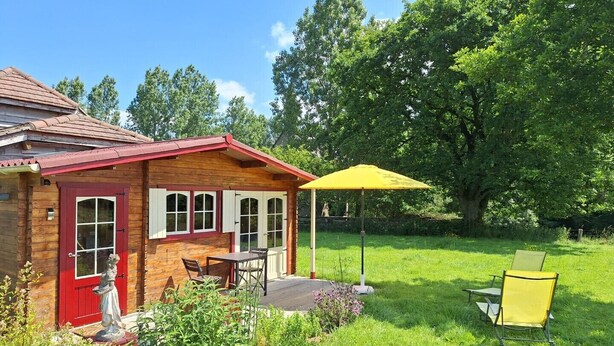 A wooden cabin with a red door, a patio with a yellow umbrella, and a statue in the foreground. - Intimate, sunny châlet for 2 people in southern Normandy (Saint-Mars-d'Égrenne)
