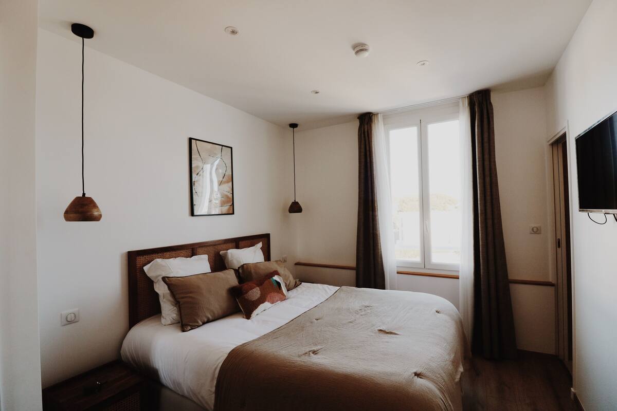 A bedroom with a large bed, two pendant lights, a framed picture on the wall, and a window with curtains.