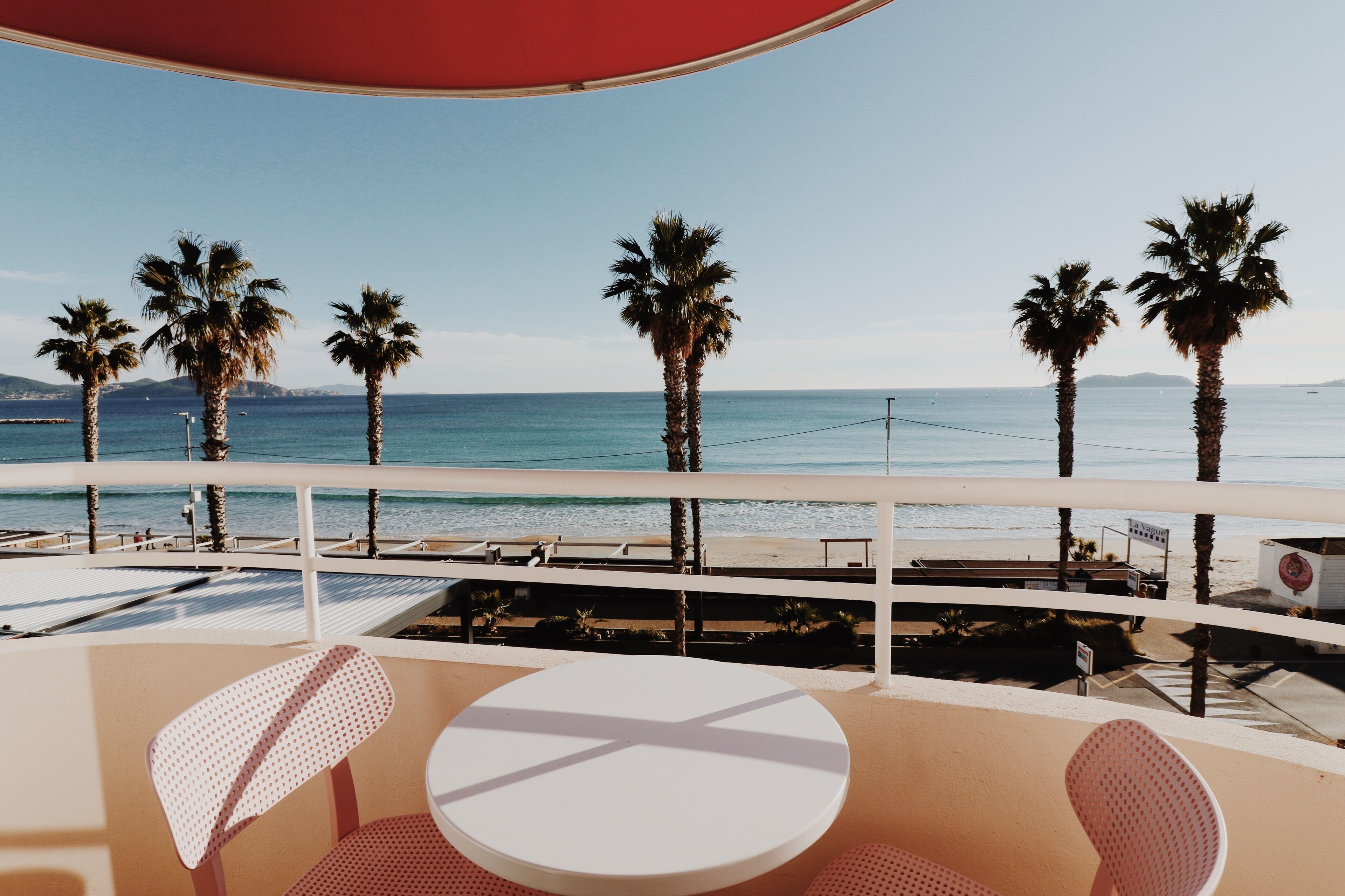 A balcony with a white table and two chairs, overlooking a beach with palm trees and the sea.