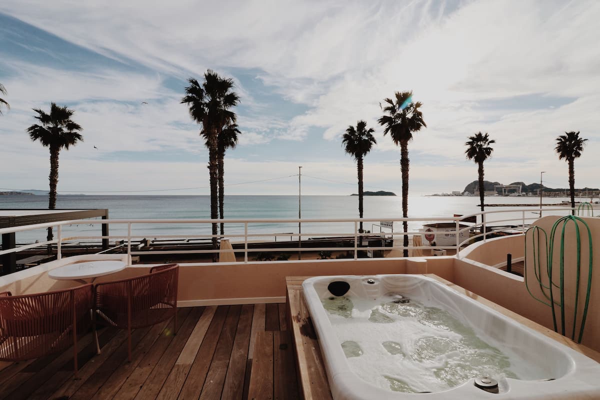 A wooden deck with a hot tub, lounge chairs, and a view of the ocean and palm trees.