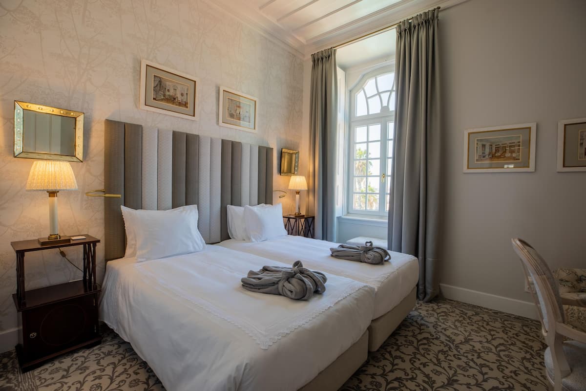 Ein Hotelzimmer mit zwei Betten, einem großen Fenster mit Vorhängen, gerahmten Bildern an der Wand und einem Sessel.