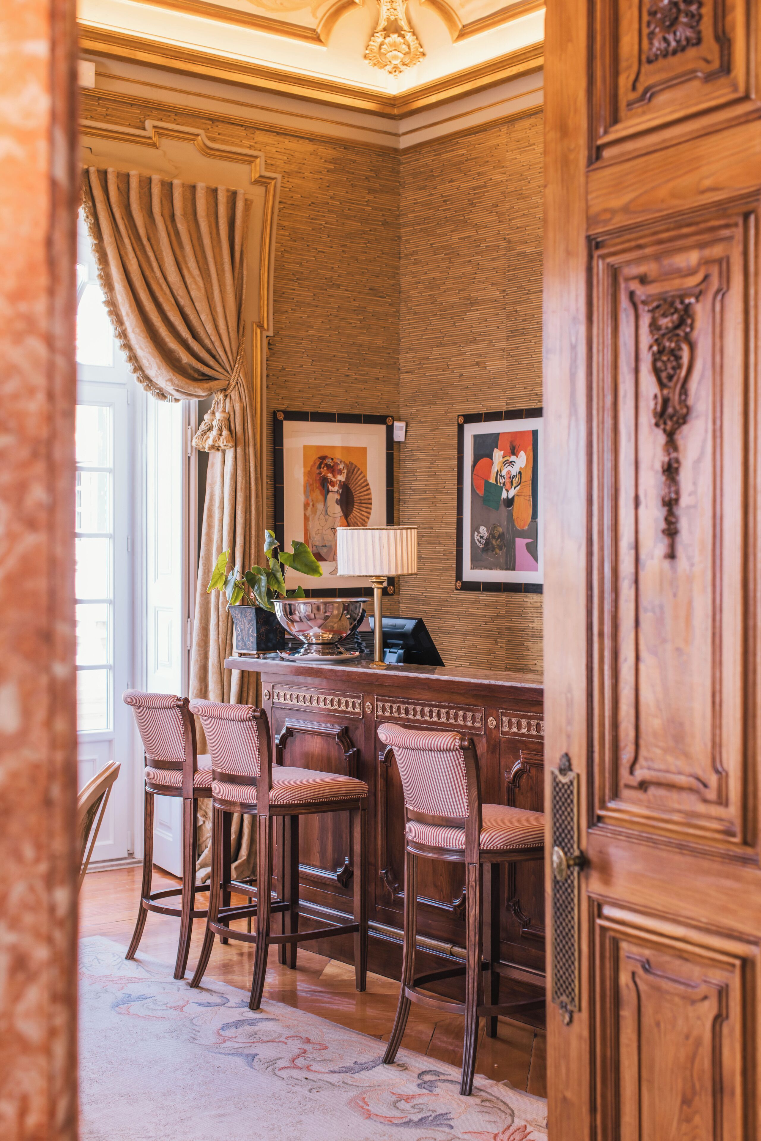 A corner room with a wooden cabinet, a desk with a lamp, and two chairs. There are two framed artworks on the wall.