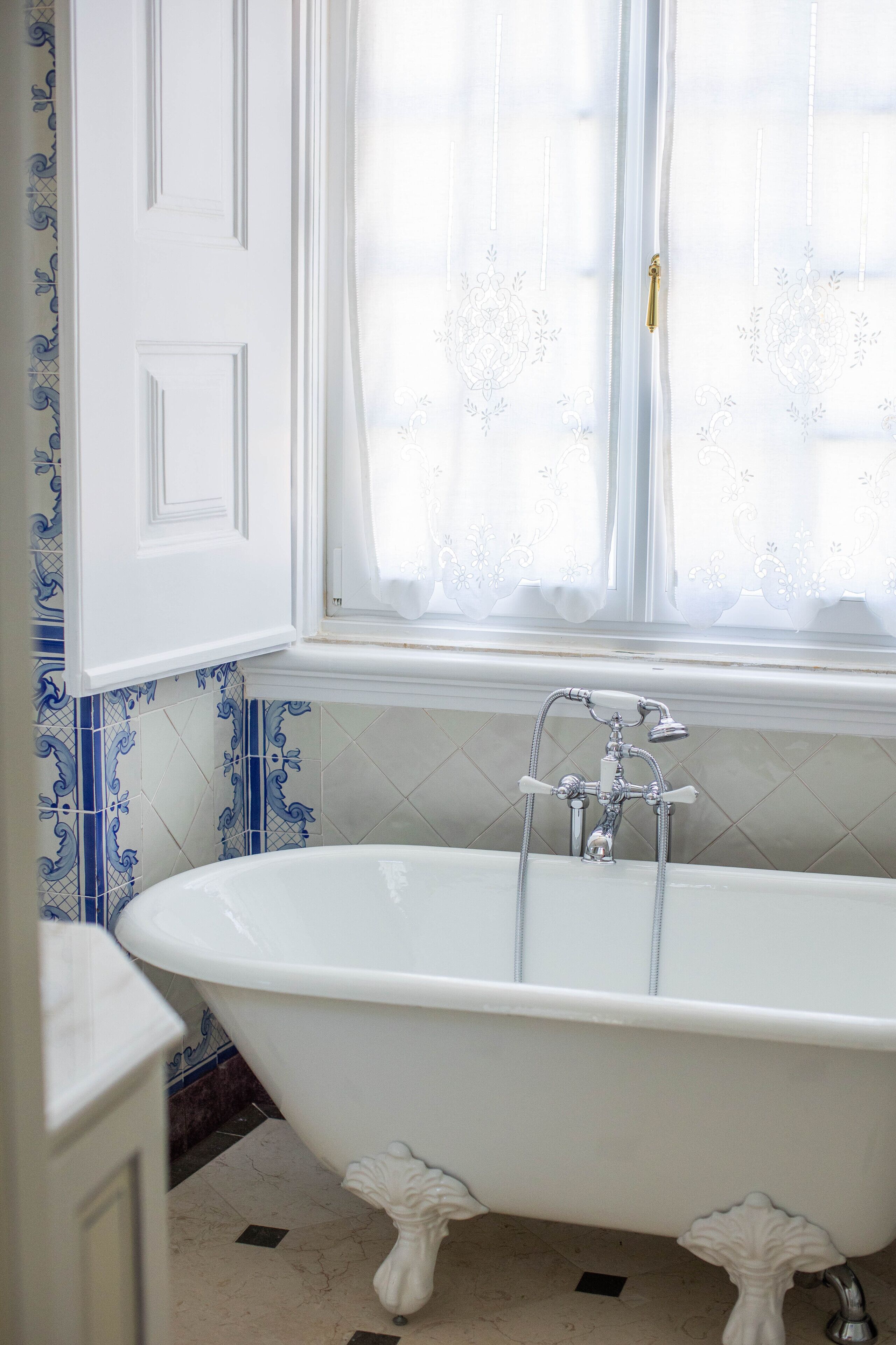 A bathroom with a freestanding bathtub, a window with curtains, and blue and white patterned tiles.