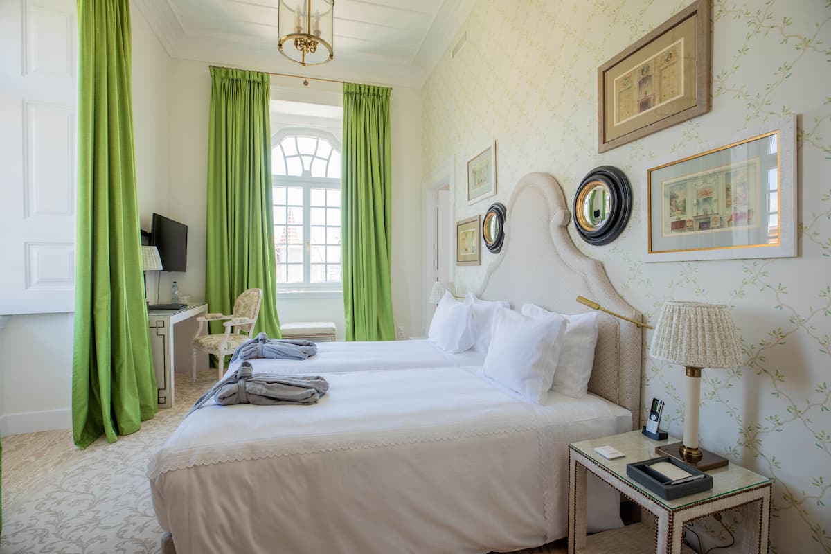 Ein Hotelzimmer mit zwei Betten, einem Schreibtisch mit Computer, einem Stuhl und einem Fenster mit grünen Vorhängen.