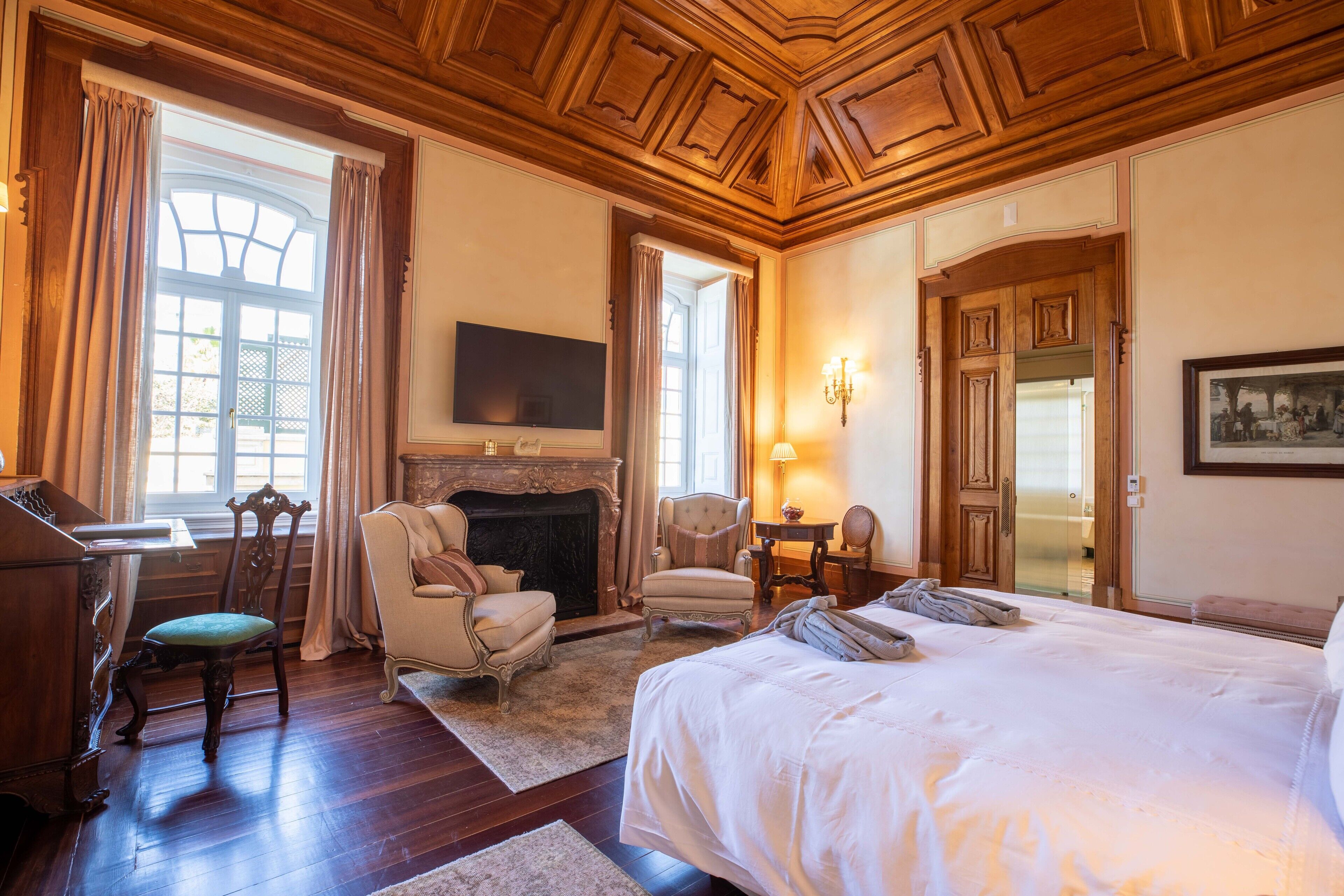 A spacious bedroom with a large bed, a fireplace, a desk with a chair, and a television mounted on the wall.