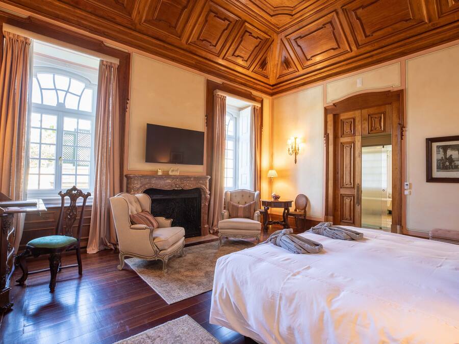 A spacious bedroom with a large bed, a fireplace, a desk with a chair, and a television mounted on the wall.