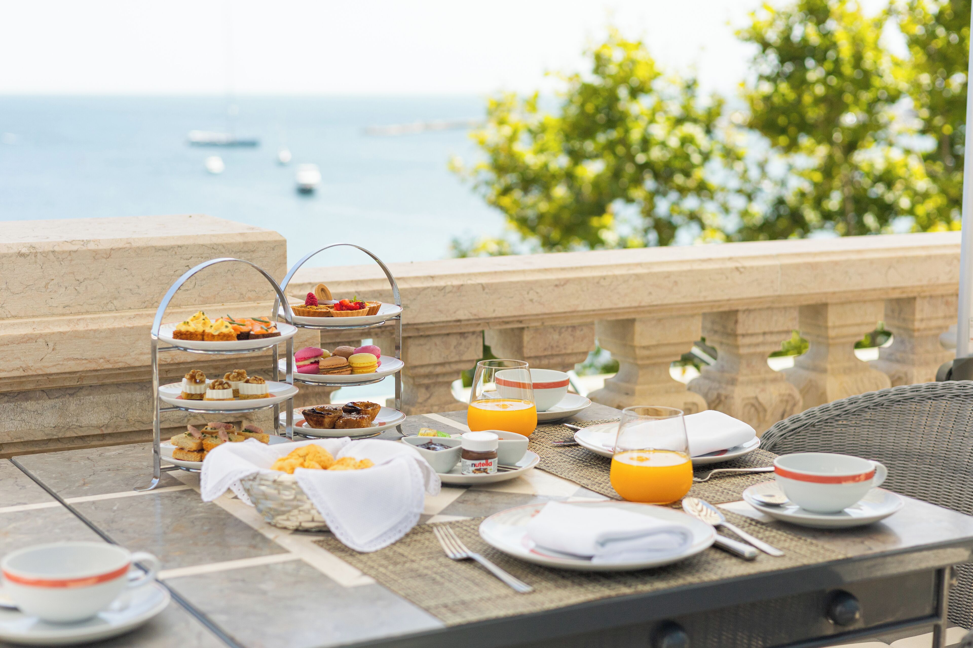 A breakfast buffet with various pastries and fruits, a glass of orange juice, and a scenic view of the sea and greenery.