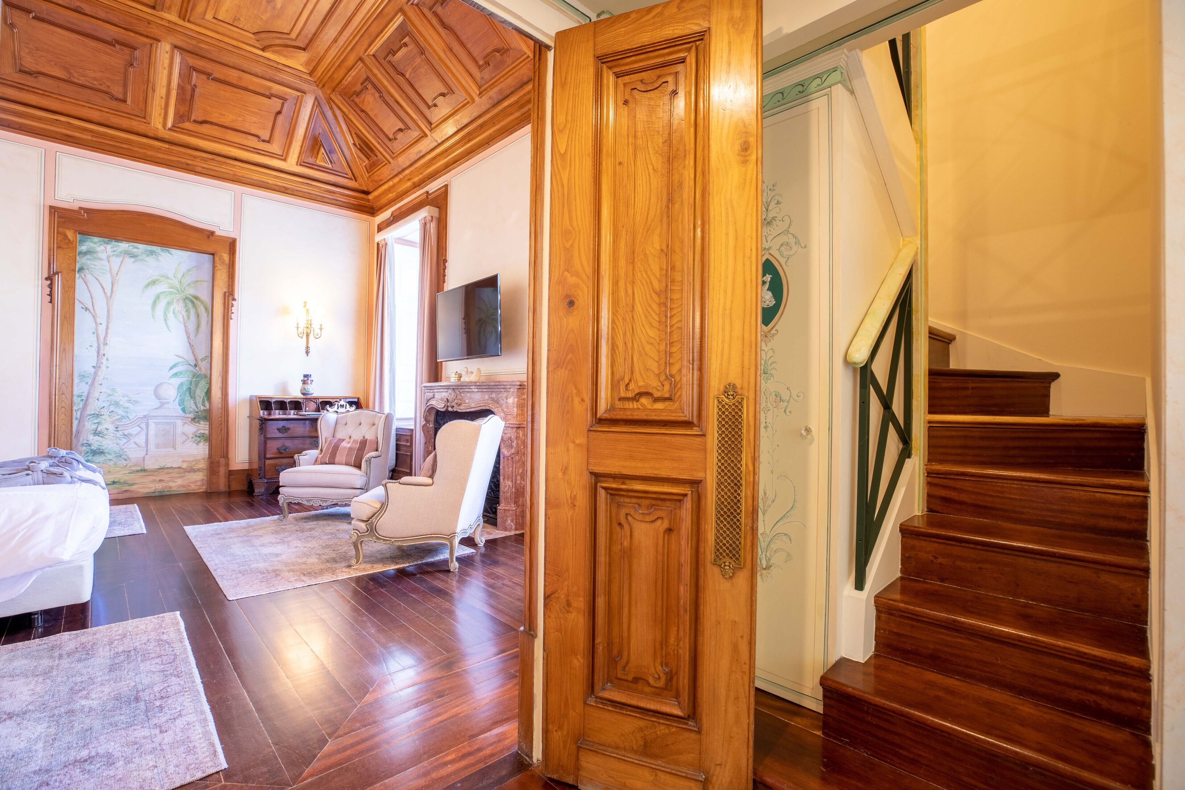 A spacious bedroom with a large bed, a sitting area with a chair and a small table, a television, and a wooden staircase.
