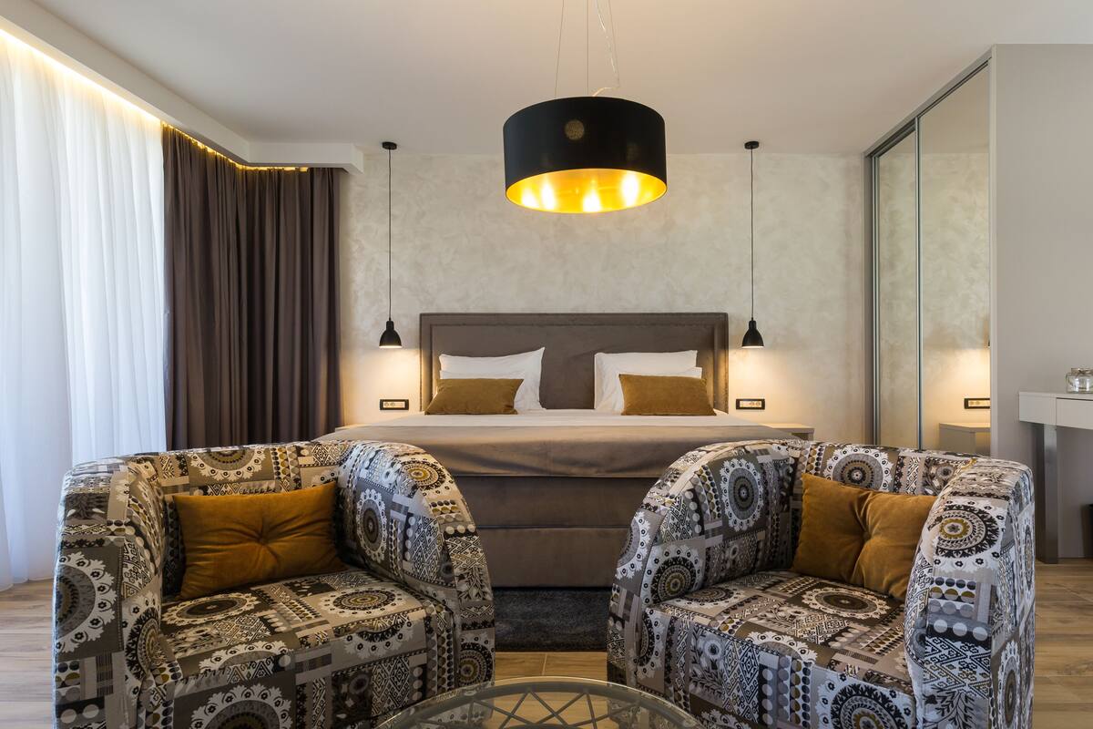A modern hotel room with a large bed, two patterned armchairs, a glass coffee table, and a pendant light above the bed.