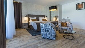 Una habitación de hotel moderna con una cama, dos sillones estampados, una mesita, una mesita de noche, un espejo y una lámpara colgante.