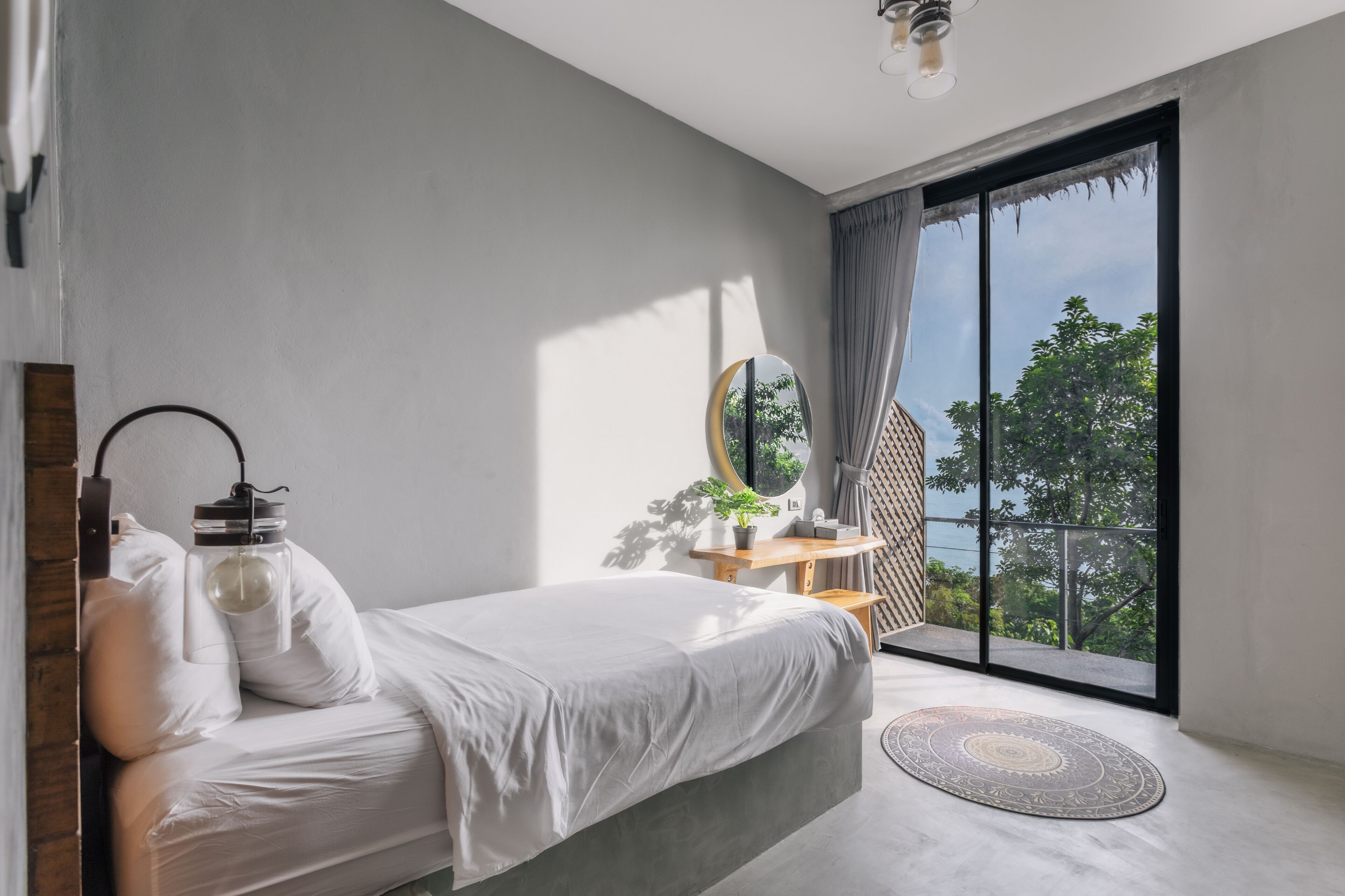 A bedroom with a bed, a bedside table with a lamp, a mirror, and a view of the outdoors through a large window.
