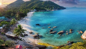 Un complejo turístico frente al mar con aguas turquesas cristalinas, una playa de arena y colinas verdes exuberantes al fondo.