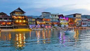 Hotel frente al agua con varias construcciones, un barco atracado y el reflejo de luces en el agua.