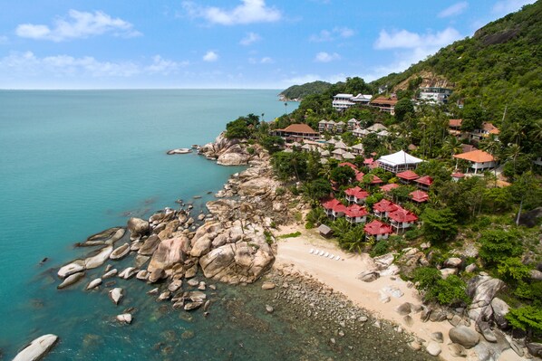 A coastal resort with a rocky beach, clear turquoise waters, and a backdrop of lush greenery and hills. - Vikasa Yoga Retreat (Koh Samui)