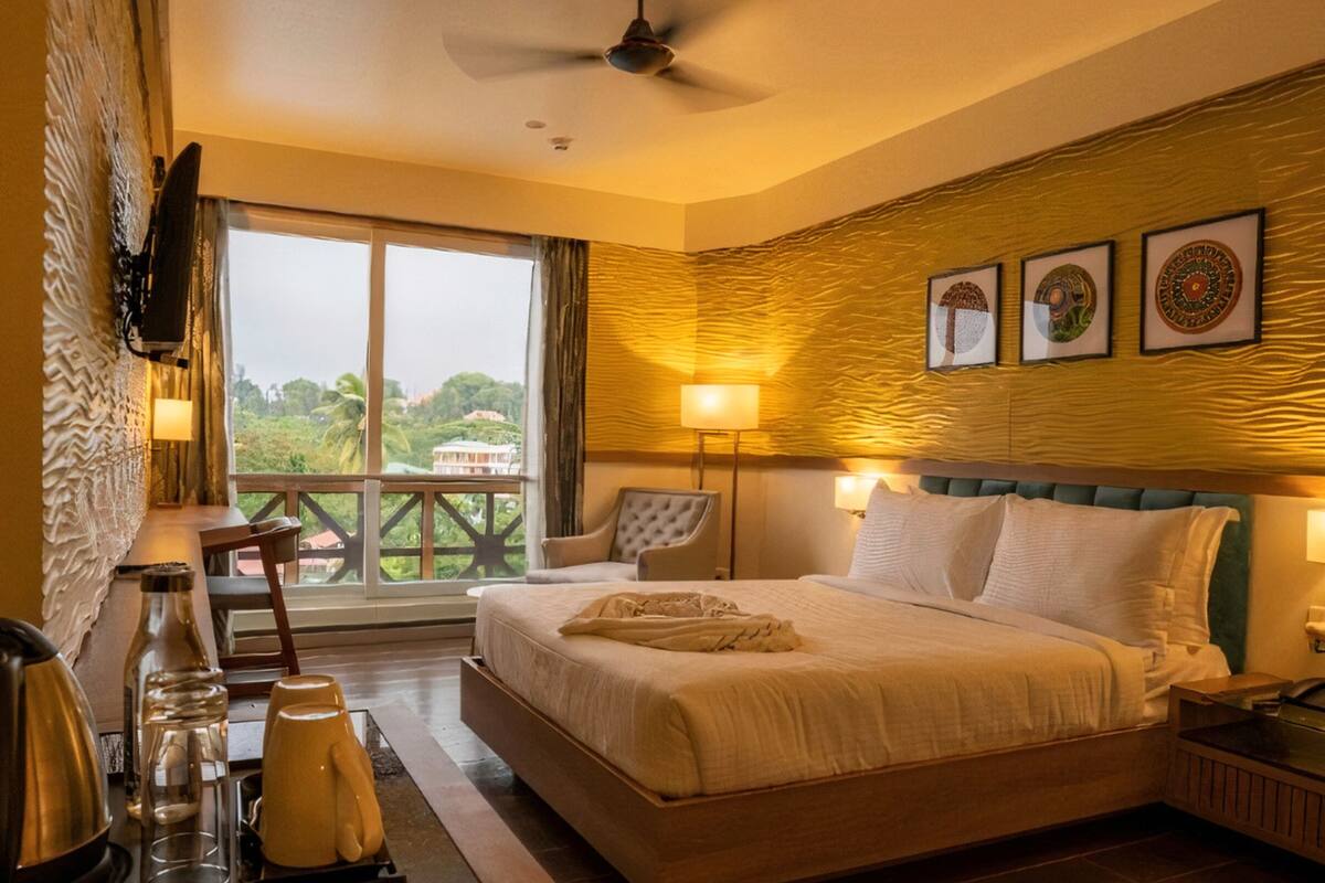 A hotel room with a bed, bedside lamps, a ceiling fan, a view of greenery outside, and a minibar.