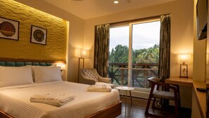 A hotel room with a large bed, a desk, two chairs, and a view of greenery through the window. - Bayleaf Inn (Port Blair)