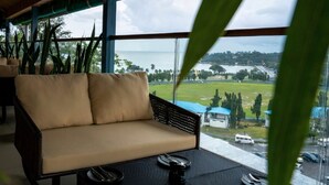 A room with a sofa and dining table, large windows with a view of a green landscape and buildings. - Bayleaf Inn (Port Blair)