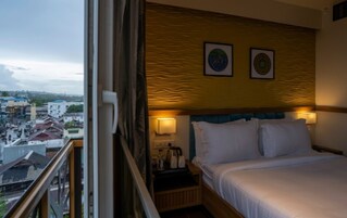 A hotel room with a bed, two wall-mounted lamps, a nightstand with a kettle, and a view of the city through a window.