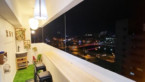 Ein Balkon mit Blick auf eine nächtliche Stadtlandschaft, mit Geländer, einer Hängeleuchte und einer kleinen Sitzecke mit Tisch und Stühlen.