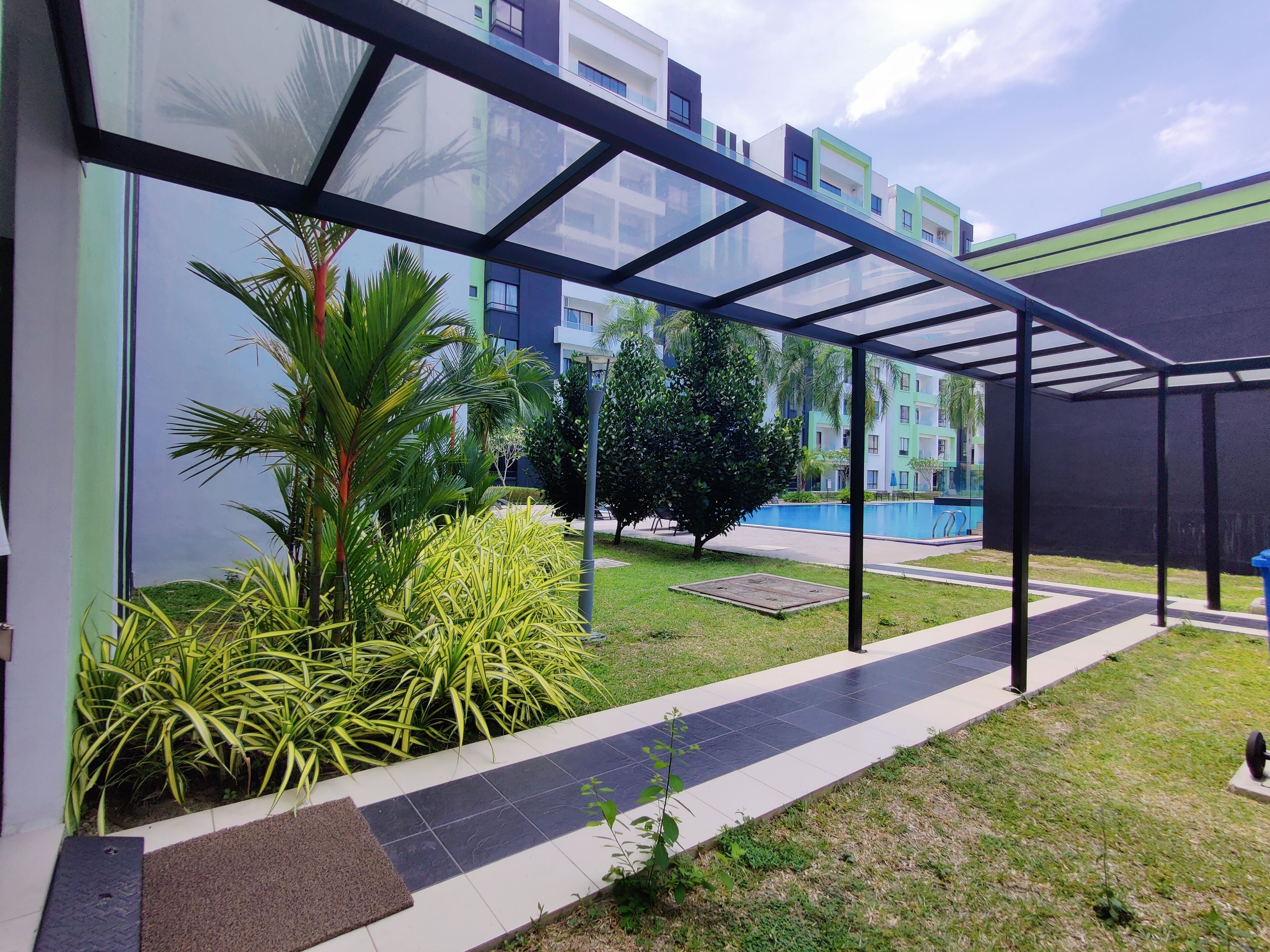 A modern outdoor area with a glass walkway, greenery, and a swimming pool in the background.