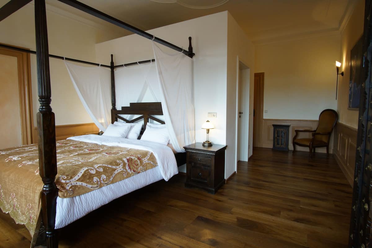 Een bed met baldakijn, een nachtkastje met lamp en een stoel in een kamer met houten vloer.