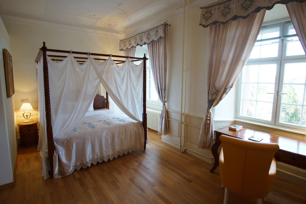 Een kamer met een hemelbed, een houten bureau en een stoel, met houten vloeren en witte muren.