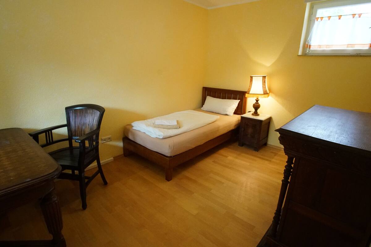 Een kleine, netjes ingerichte slaapkamer met een enkel bed, een stoel, een tafel en een nachtkastje met een lamp.