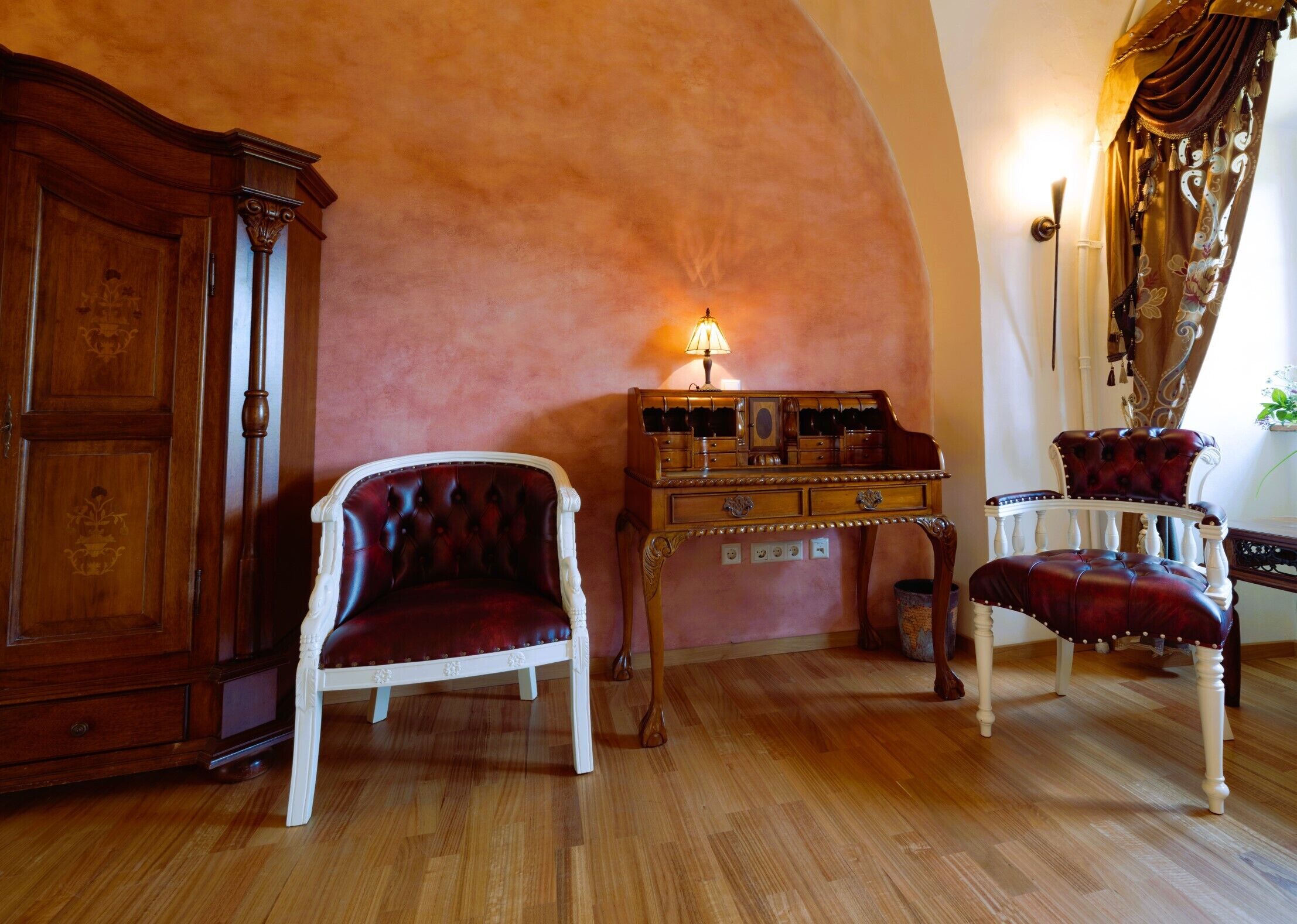 A room with a wooden cabinet, a tufted armchair, a small table with drawers, and a lamp.