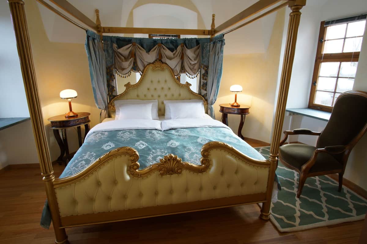 Een hemelbed met een geborduurde hoofdbord, twee nachtkastjes met lampen, een stoel en een raam met gordijnen.