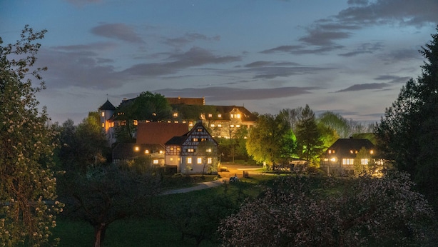 A large, illuminated estate with multiple buildings, surrounded by lush greenery and flowering trees.