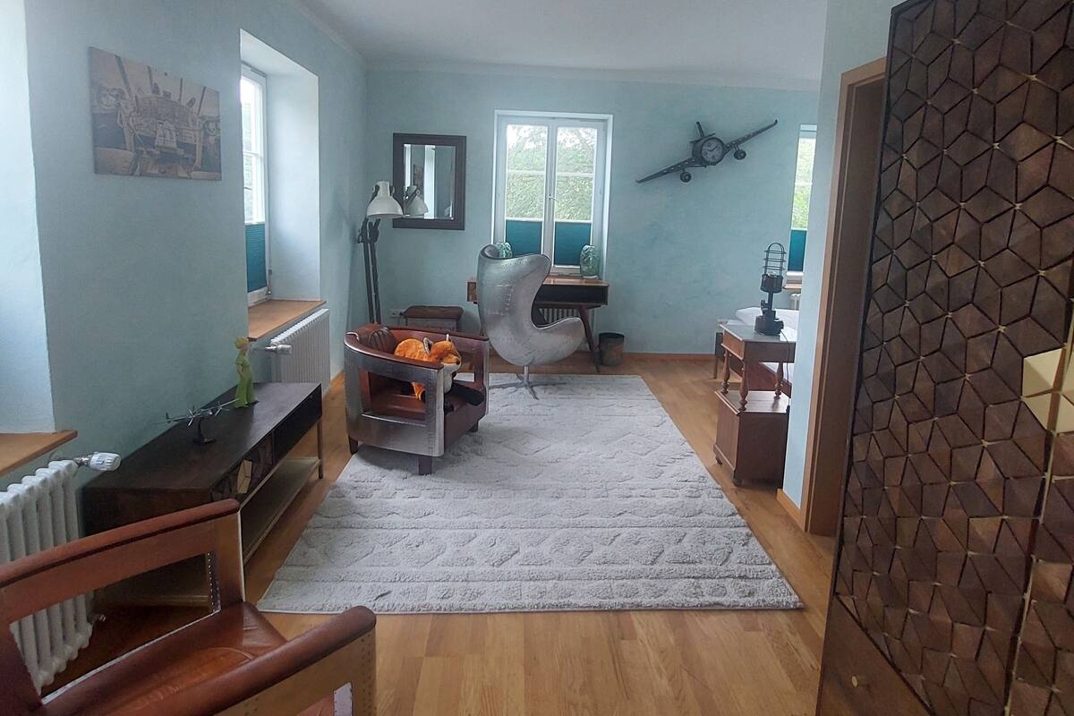 Een ruime woonkamer met een groot raam, een plafondventilator en een decoratief wandpaneel.