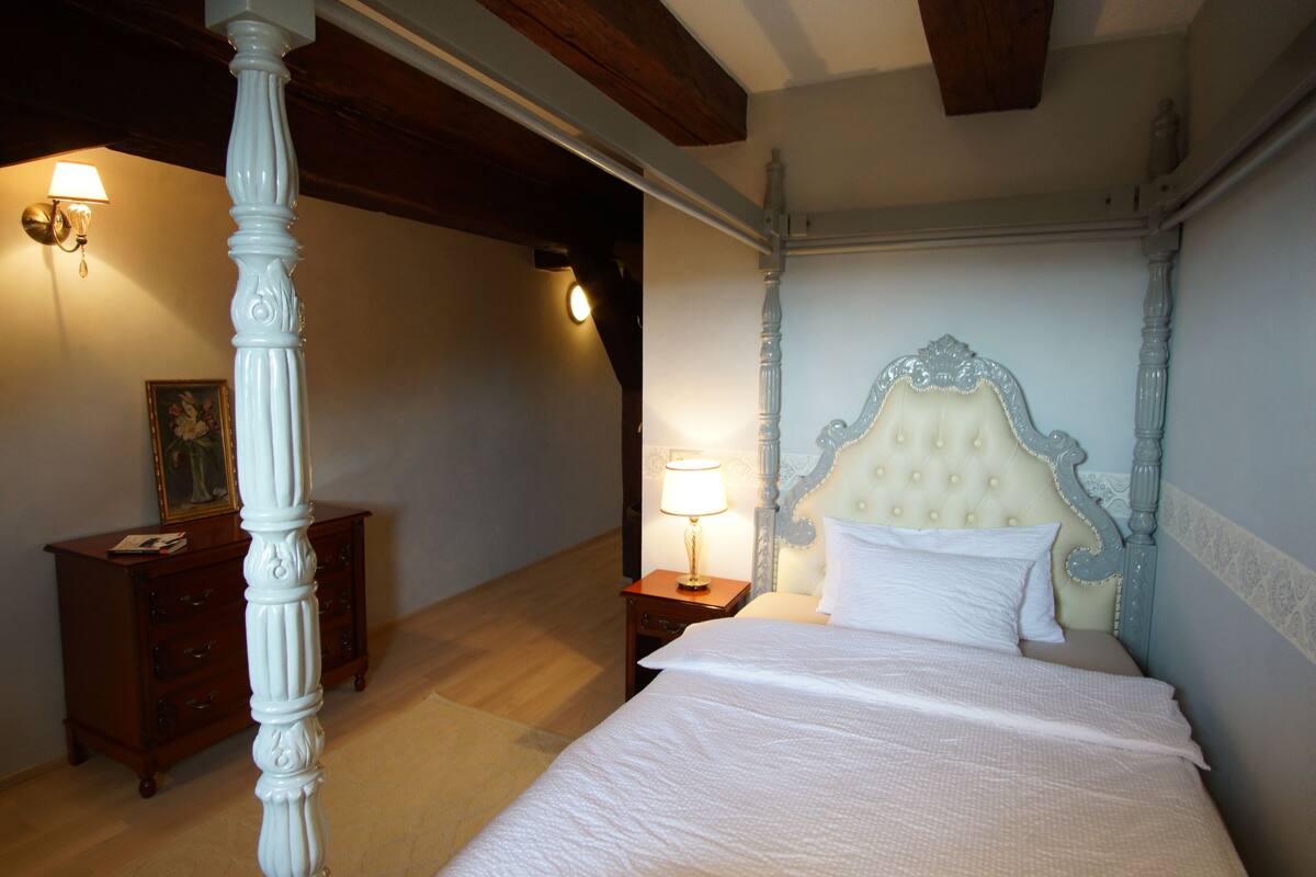 Een slaapkamer met een hemelbed, een nachtkastje met een lamp, een kaptafel en een ingelijste foto op de kaptafel.