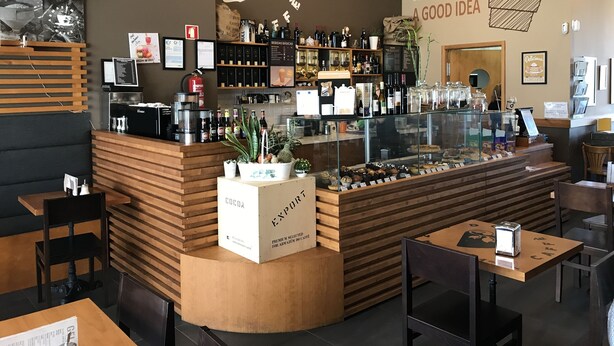 Interno di un bar con un bancone in legno, scaffali di chicchi di caffè e bottiglie, una vetrina con dolci e una parete con un'illustrazione di una tazza di caffè e del testo.