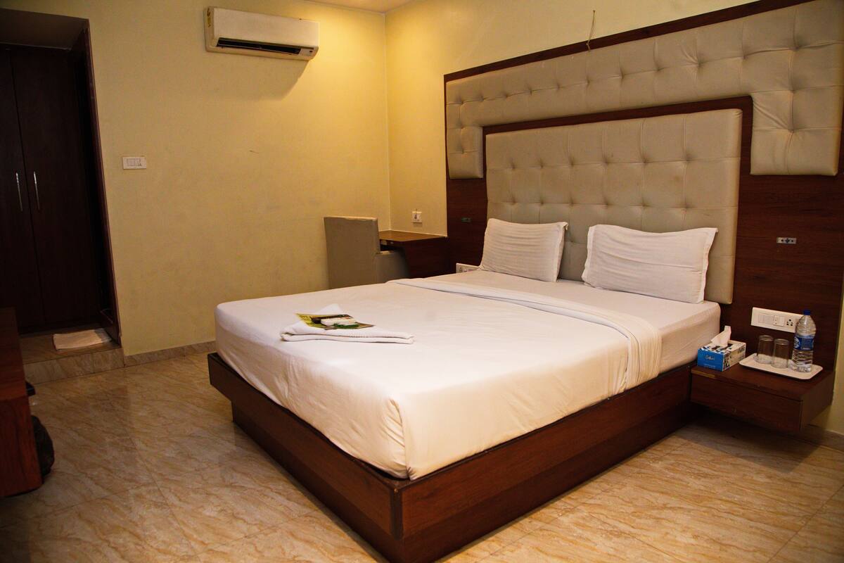 Une chambre d’hôtel avec un grand lit, une tête de lit en bois, une table de chevet avec une bouteille et des mouchoirs, et un appareil de climatisation fixé au mur.