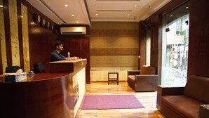 A reception area with a wooden reception desk, a small table, a sofa, and a large window. - Hotel Dalon (Mumbai)