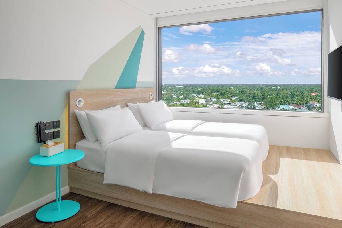 A modern hotel room with a bed, a small table, and a large window offering a city view.