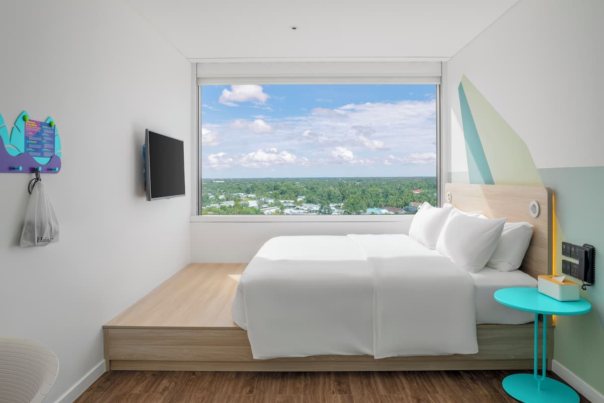 A modern hotel room with a large window, a bed with white linens, a small round table with a lamp, and a wall-mounted TV.