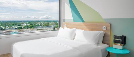 A modern hotel room with a large bed, a view of the city, and a small table with a phone and a tissue box.