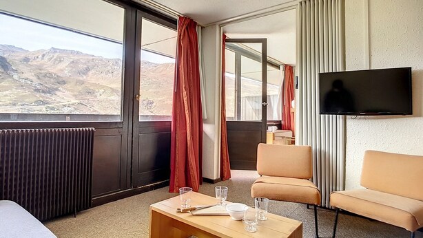 A hotel room with a large window offering a mountain view, a flat-screen TV mounted on the wall, a wooden coffee table with glasses and a book, and two armchairs. - Bright apartment for 6, ski-in/ski-out, south balcony (Les Menuires)