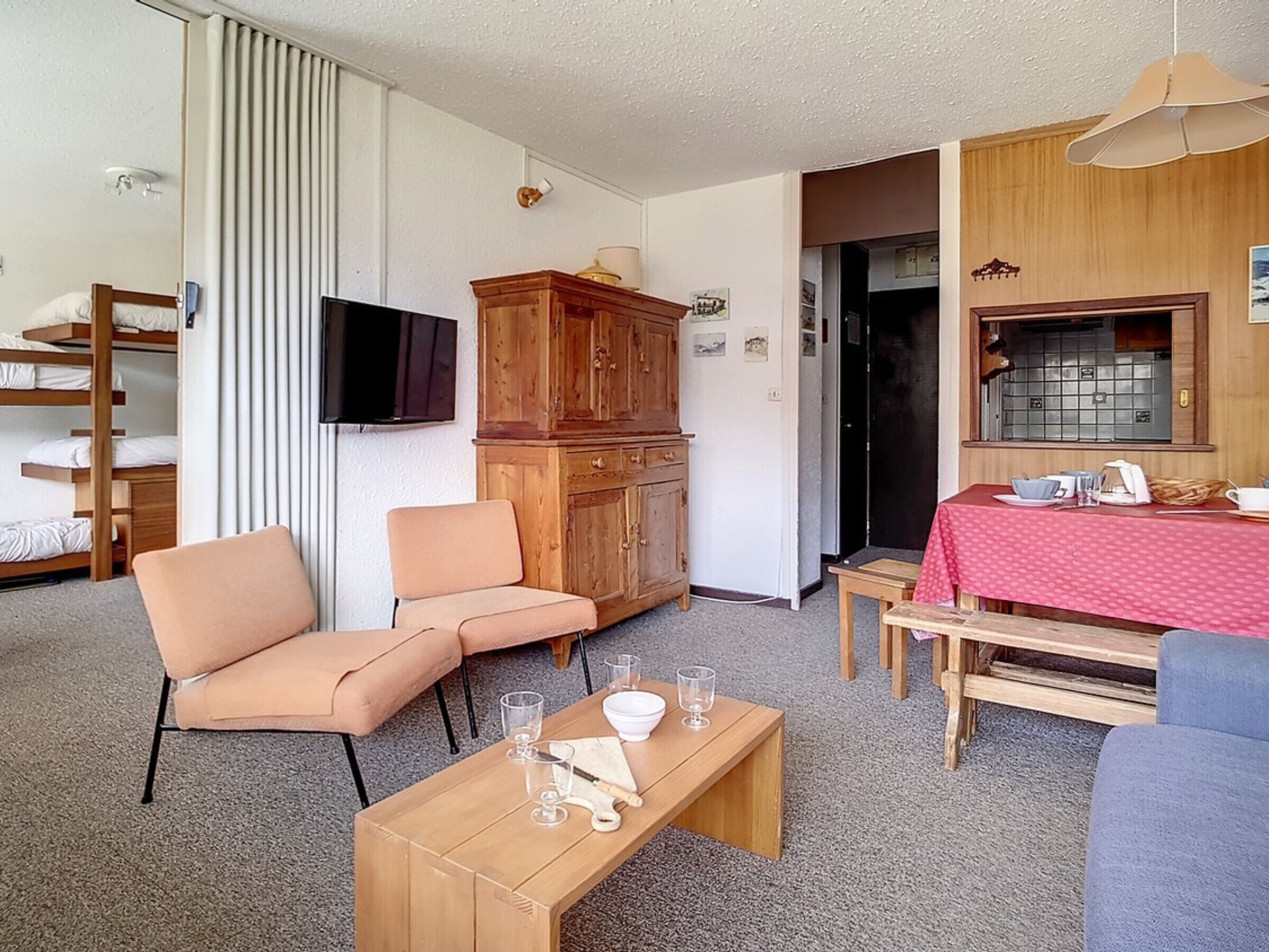 Un espacio habitable compacto con sofá, una mesa de madera, televisor y una cocina con mantel rojo, además de un mueble de madera.