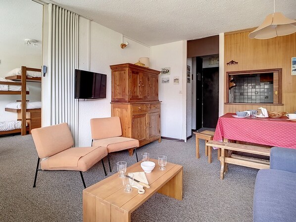 Un espacio habitable compacto con sofá, una mesa de madera, televisor y una cocina con mantel rojo, además de un mueble de madera.