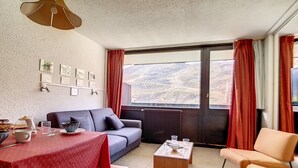 A cozy living area with a sofa, a dining table with chairs, a coffee table, and a view of a mountainous landscape through a large window. - Bright apartment for 6, ski-in/ski-out, south balcony (Les Menuires)