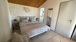 Um quarto com teto de madeira, uma cama com roupa de cama estampada, uma mesa de cabeceira com abajur e uma cadeira.