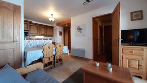 Ein gemütliches Wohnzimmer mit Bett, einem Holzesstisch und einem Fernseher. Es gibt eine Küchenzeile mit Holzschränken und einer Mikrowelle.