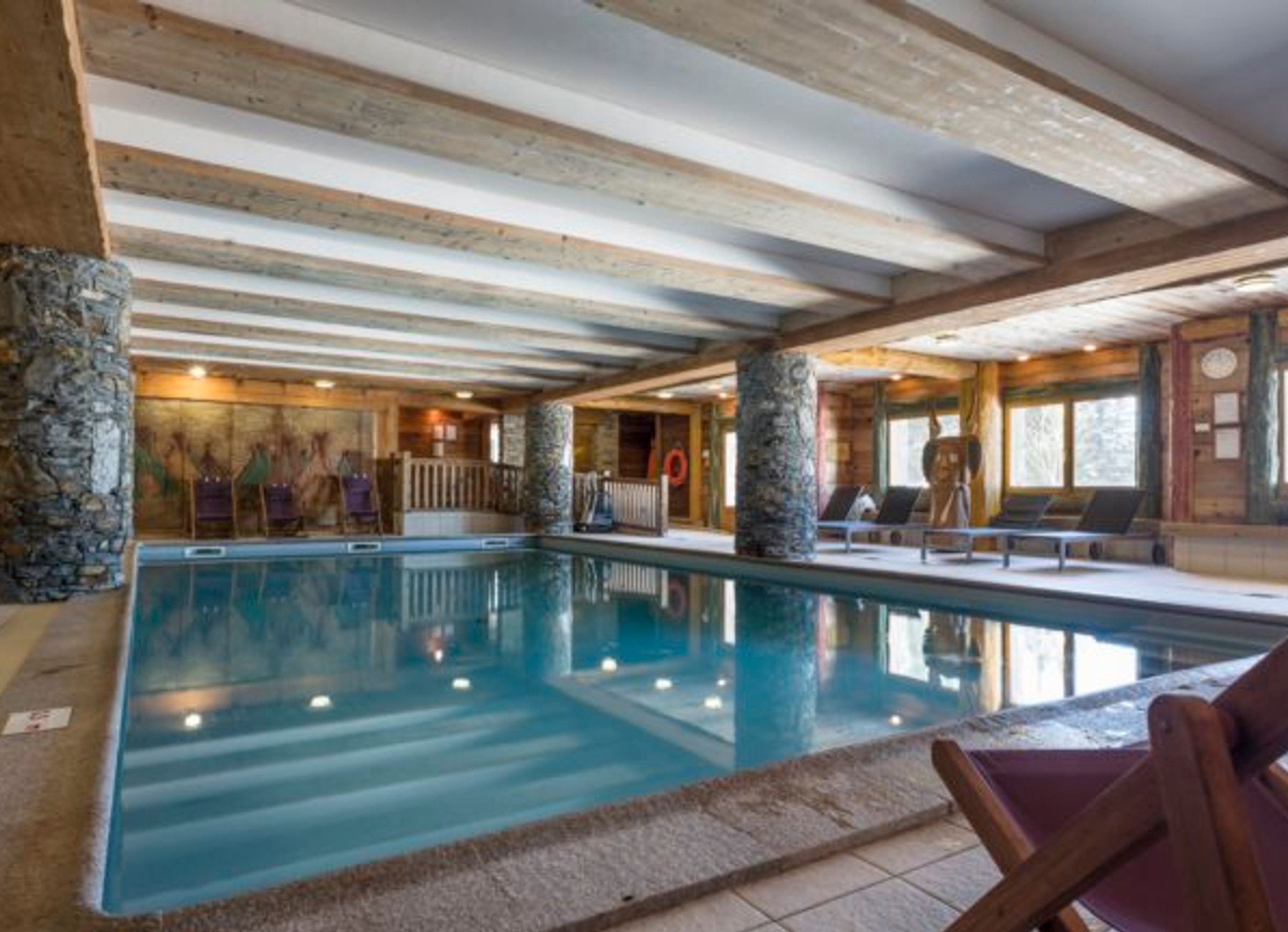 Une piscine intérieure agrémentée de piliers en pierre, de poutres en bois et d’un mur en pierre.