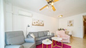 Ein Wohnzimmer mit grauem Sofa, einem kleinen runden Tisch, einem rosa Teppich und einem Deckenventilator.
