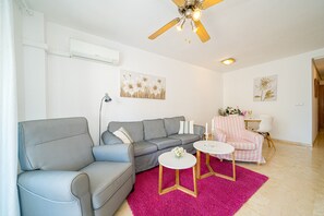 Ein Wohnzimmer mit grauem Sofa, einem kleinen runden Tisch, einem rosa Teppich und einem Deckenventilator.